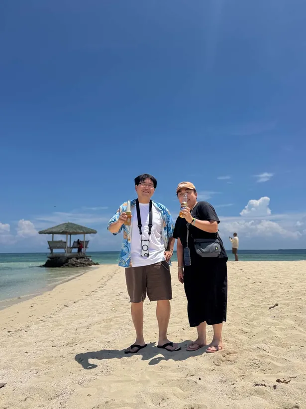 パンダノン島の砂浜の先に立つ東屋を背景にビールで乾杯する2名様
